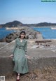 A woman in a green dress leaning against a wall by the water.